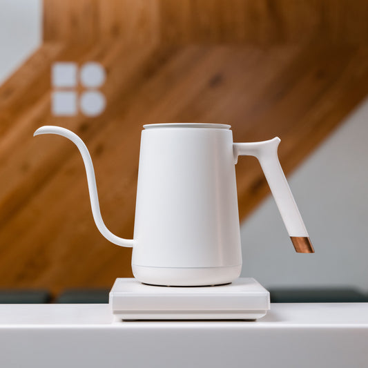 white timemore pour over electric kettle on a white table and wooden back drop