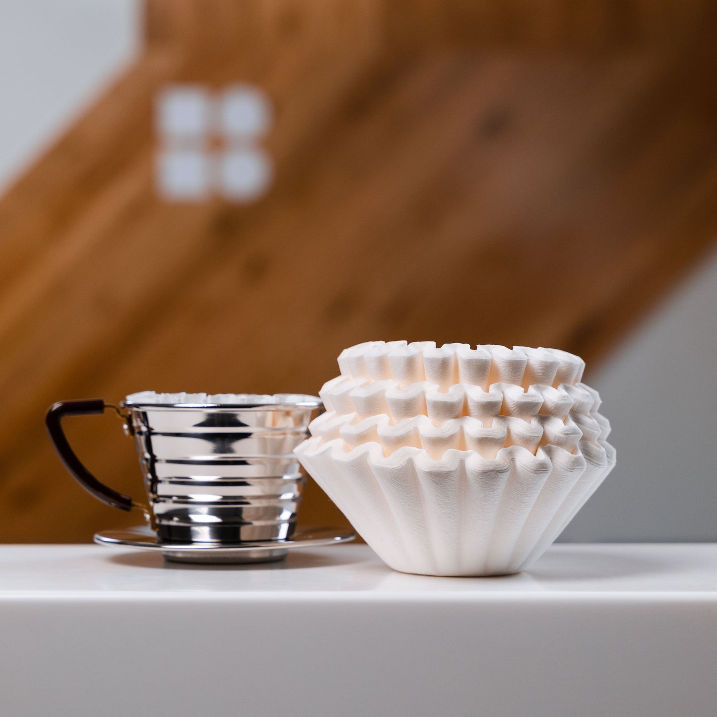 Kalita wave filter papers, beside a stainless steel kalita dripper, on a white surface against a wooden backdrop
