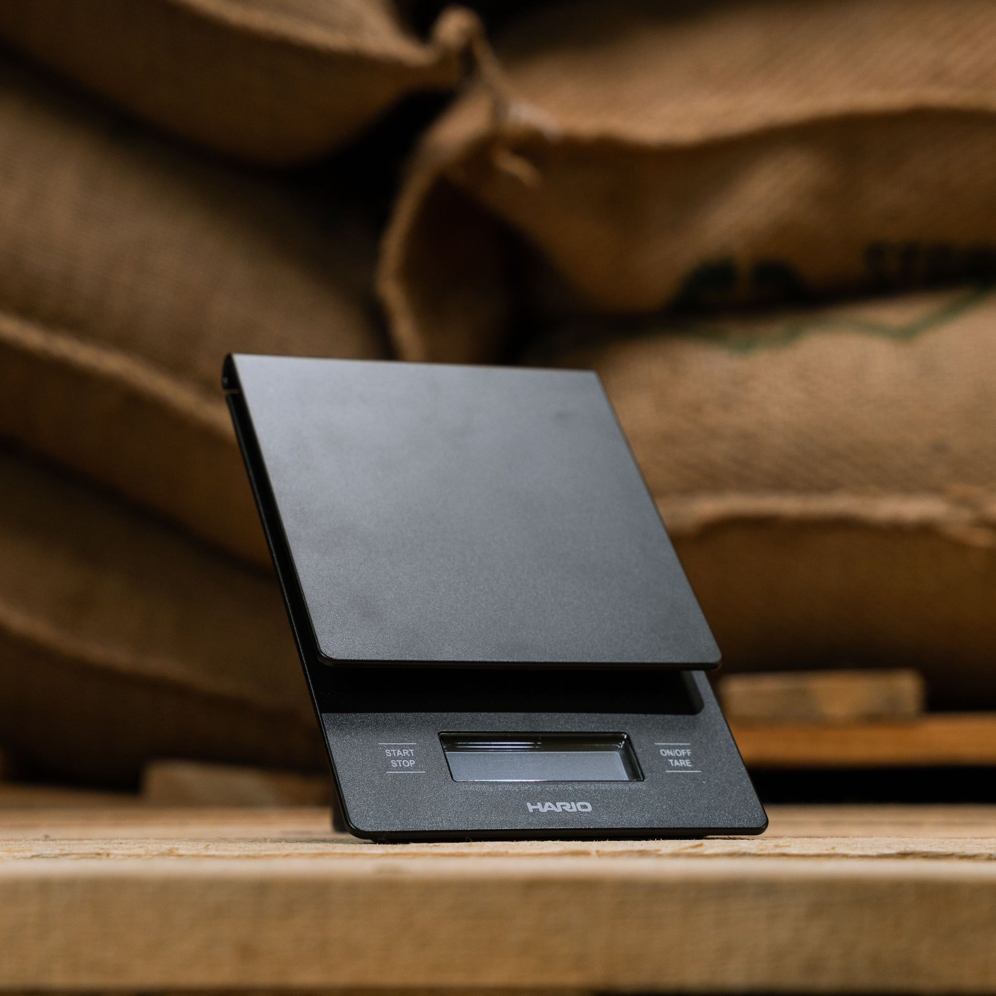 hario drip scales displayed at a 45 degree angle and in front of green bean coffee bags
