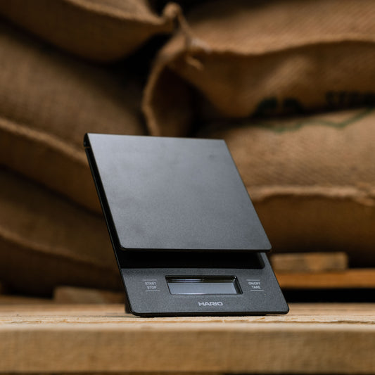 hario drip scales displayed at a 45 degree angle and in front of green bean coffee bags
